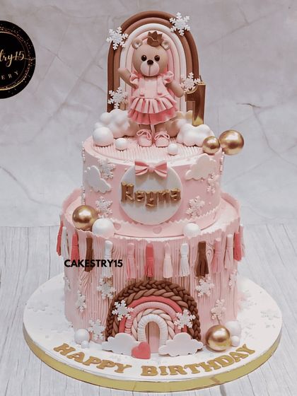 A dreamy two-tier teddy bear cake for a first birthday. This 3kg chocolate cake features a soft pink color, a teddy on a rainbow, and delicate snowflake accents.