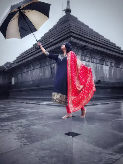 A striking color pop photograph. The woman's vibrant red dupatta stands out against the monochrome background of a traditional temple on a rainy day, creating a visually powerful image.