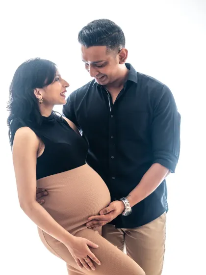 A bright and happy studio portrait against a clean white background. The couple's smiles and the dad-to-be's gentle touch on the bump radiate pure joy.