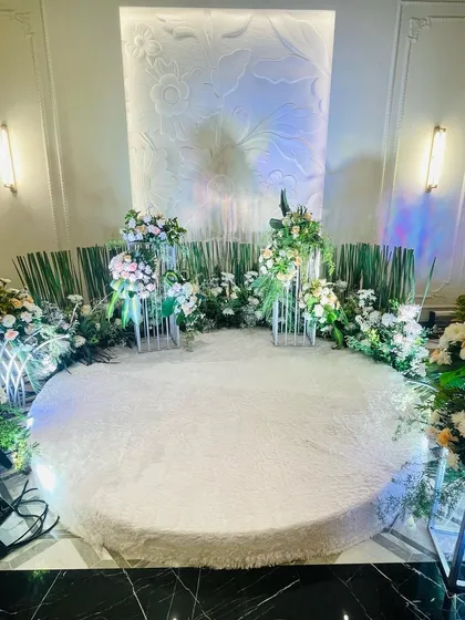 A close-up of the circular stage for an all-white engagement ceremony. The plush white carpet and surrounding floral arrangements create a soft and luxurious feel.