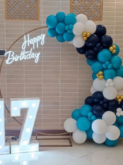 A cool and stylish 17th birthday setup. The semi-arch of blue, teal, and white balloons against a circular frame with a neon sign and light-up numbers is perfect for a teen's party.