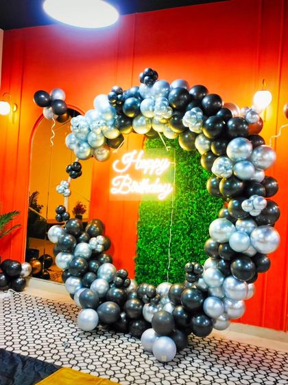 A bold and stylish ring decoration with black and silver balloons against a vibrant orange wall and a green hedge backdrop. The neon sign adds a cool, modern touch.