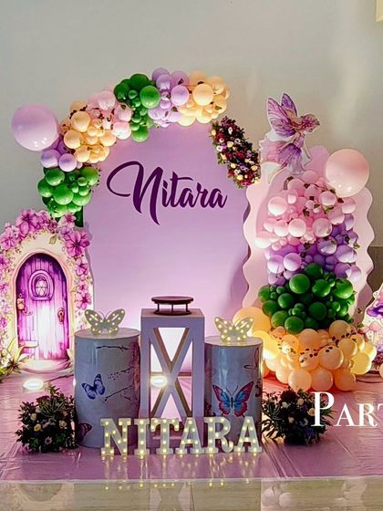 A whimsical fairy theme party with a magical door prop. The purple backdrop is complemented by colorful balloon arrangements, butterfly-themed pedestals, and light-up marquee letters.
