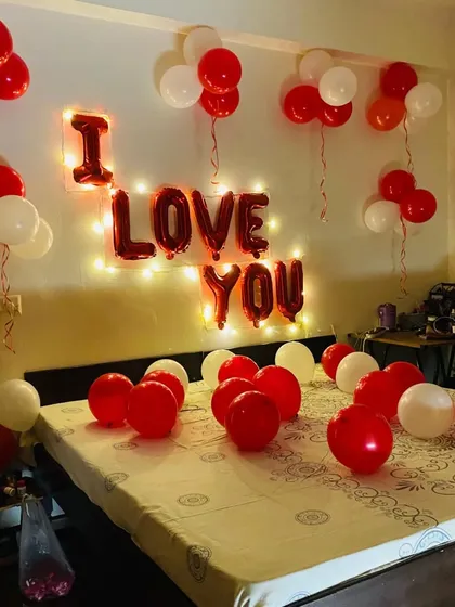 A classic 'I LOVE YOU' room decoration. The red foil letters are backlit with fairy lights for a warm glow, and the room is filled with red and white balloons on the bed and hanging from the ceiling.