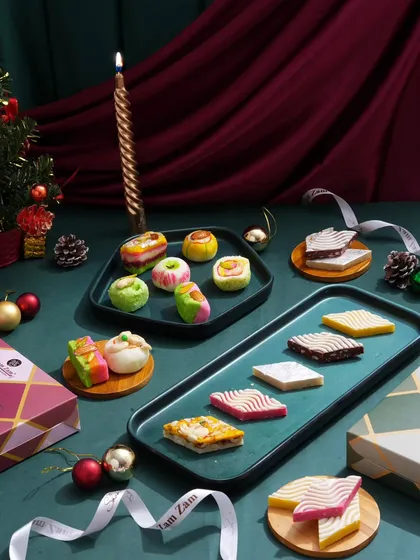 This shot captures a sophisticated spread of Indian sweets styled for a Christmas campaign. The deep green and red tones, along with the candlelight, create a warm and festive atmosphere.