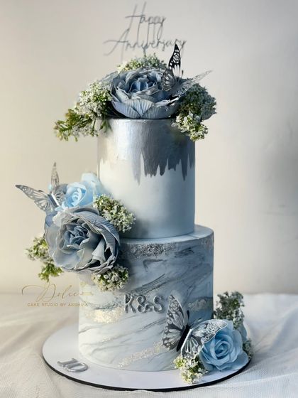 A beautiful two-tiered cake for an anniversary celebration. The bottom tier has a grey and silver marble effect, while the top has a silver lustre finish, all adorned with blue sugar flowers.