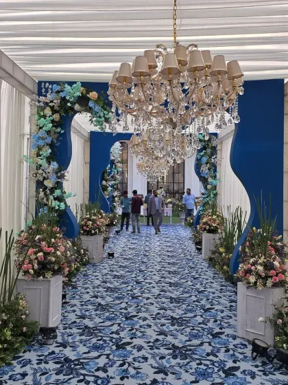 A grand entrance for Mehak and Hem's event, featuring a blue and white floral-printed carpet, chandeliers, and dramatic blue walls framed by floral arches.