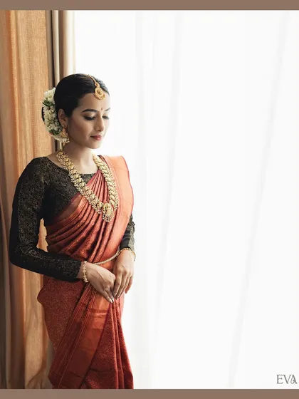 A serene, full-length portrait of the model standing by a window. The soft, natural light beautifully illuminates the texture of the saree and her graceful posture.