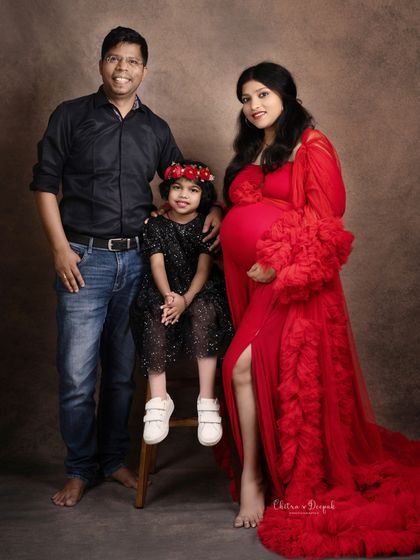 A classic family maternity photo with the expecting mother in a vibrant red gown, standing with her husband and daughter.