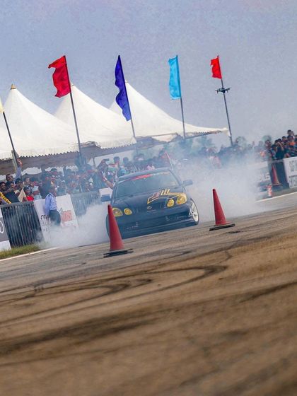 A drift car slides past the crowd, flags waving in the background. The combination of speed, smoke, and sound creates an electric atmosphere.