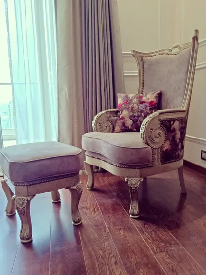 A close-up of a custom statement armchair, perfect for a quiet reading corner. This piece features a high back, ornate carved wooden frame, and a combination of solid and floral upholstery, complete with a matching footstool for ultimate comfort.
