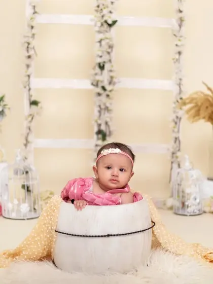 A sweet baby girl in a pink dress peeks out from a white bucket. The simple, bright background with floral accents creates a clean and charming portrait.