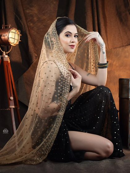 A seated portrait where the model is wrapped in a sheer, gold-embroidered dupatta. The studio props and warm lighting enhance the traditional yet modern feel of the image.