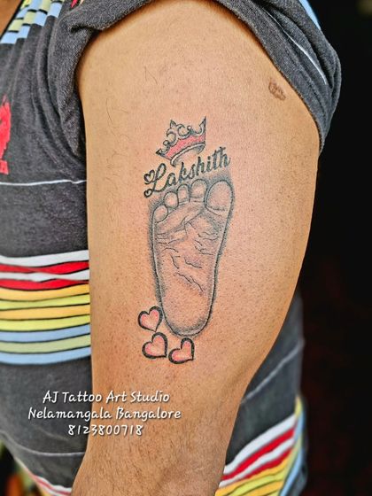 A precious memory made permanent. This tattoo features a baby's footprint, name, and a small crown, a perfect tribute for a new parent on their upper arm.