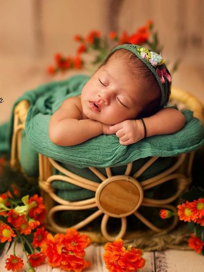 A newborn rests peacefully in a floral-detailed basket, wrapped in a green swaddle and surrounded by bright orange flowers. The color contrast creates a vibrant and lively portrait.