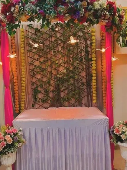 A rustic yet festive mandap design featuring a bamboo lattice backdrop intertwined with green vines and delicate bird-shaped lights. The frame is draped in bright pink fabric and decorated with marigold garlands for a traditional feel.