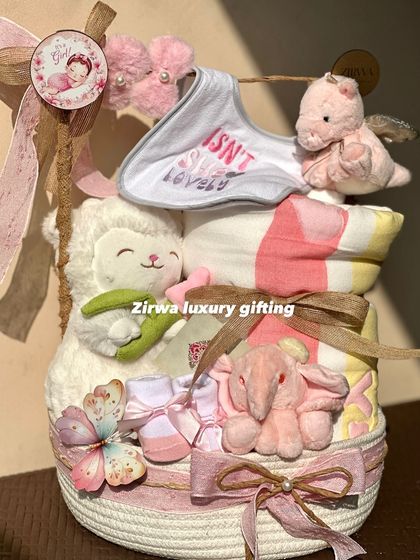 Isn't she lovely? This beautiful hamper for a baby girl is stacked with essentials, including a soft blanket, plush toys, and tiny shoes, all in a sweet pink and white color scheme.