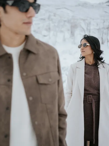 A modern, stylish shot with a focus on composition. The groom is in the foreground, slightly blurred, while the bride looks towards him from a distance, creating a dynamic and visually interesting pre-wedding portrait in the snow.