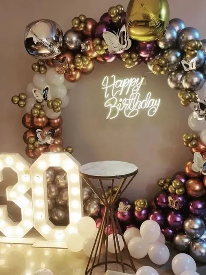 A chic 30th birthday party with a circular balloon arch in metallic colors. The setup includes a neon sign and butterfly accents for a touch of whimsy.