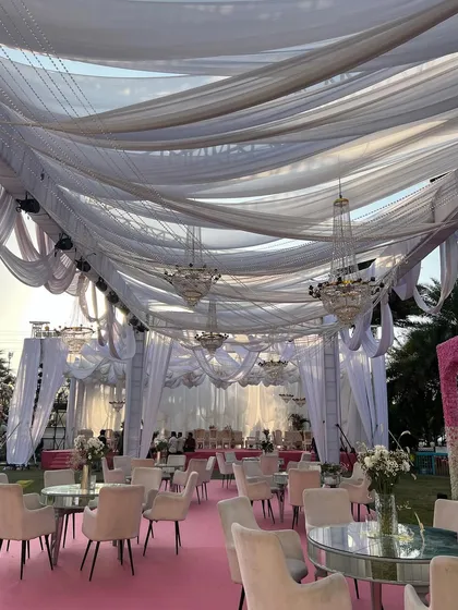 A wide shot of the outdoor cocktail setup, with its ceiling of elegant white drapes and chandeliers. The combination of soft fabrics and sparkling crystals creates a luxurious and dreamy environment for an evening celebration.