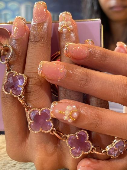 A close-up of a glamorous manicure. These nails feature a glittery gold French tip and are accented with a beautiful 3D flower made of tiny pearls.