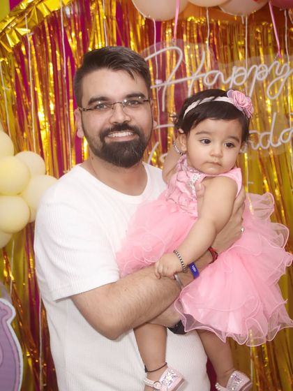A father holds his daughter in front of a pink and gold backdrop at her birthday party.