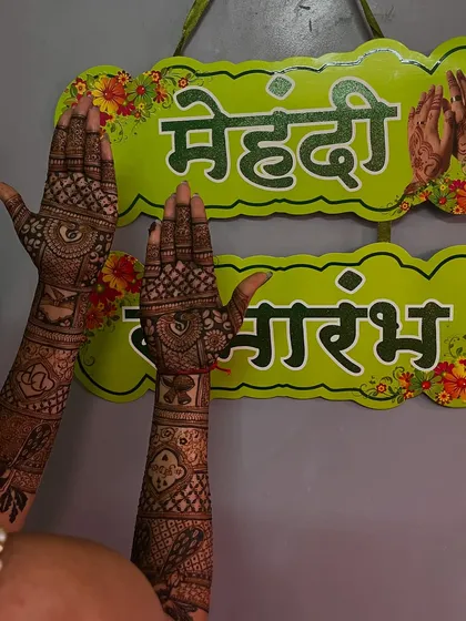 A bride posing next to a "Mehendi Samarambh" sign, her hands beautifully decorated for the ceremony.