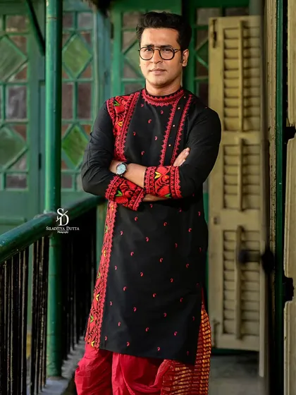 Actor Anirban Bhattacharya in a black embroidered kurta for a Pujo shoot. His confident, arms-crossed pose against the classic green slatted doors creates a strong and stylish portrait.