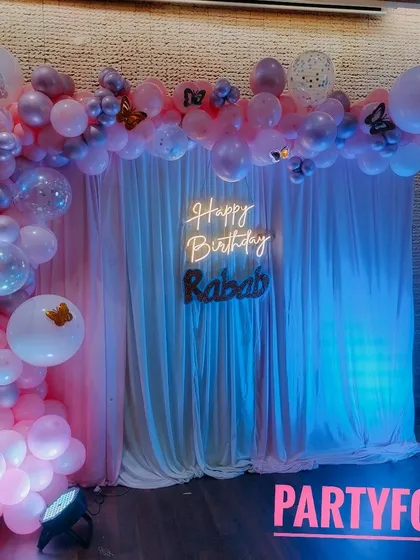 A lovely birthday decoration for Rabab, featuring a pink and purple balloon arch over a white curtain backdrop with a custom neon sign.