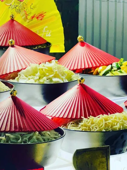 A beautifully arranged station for Chinese cuisine. Guests can choose from a variety of noodles, rice, and vegetables, all presented in thematic bowls with distinctive red fan-shaped lids.