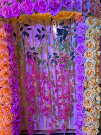 This photo shows a backdrop illuminated with colored lighting. The frame of yellow and pink artificial roses glows under the lights, highlighting the hanging floral vines in the center.