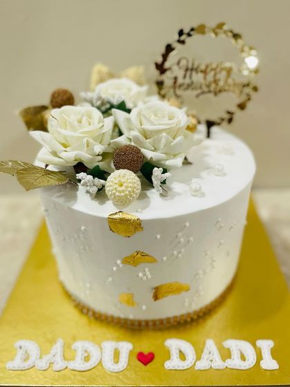 An elegant anniversary cake for "Dadu & Dadi." This classic white cake is topped with beautiful white roses and accented with gold details.