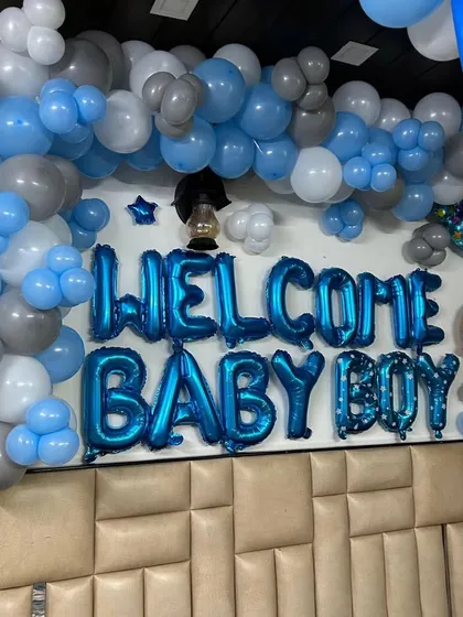 A close-up of a "Welcome Baby Boy" wall decoration. The setup features blue foil letter balloons surrounded by an arch of blue, grey, and white balloons, perfect for a bedroom or living room wall.