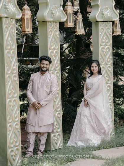 A formal portrait of a couple in elegant, coordinated traditional wear, posing between ornate pillars in a garden.