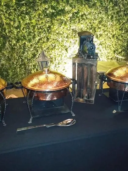 An intimate buffet arrangement against a green wall, featuring hammered copper chafing dishes on wrought iron stands and decorative props like a miniature train for a themed event.