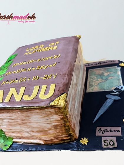 Another view of the "Lord of Numbers" book cake, personalized with the name "ANJU" in gold lettering.