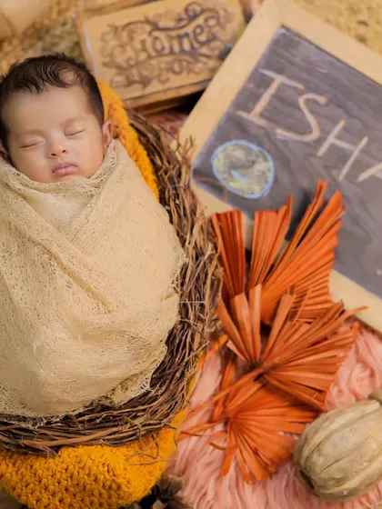A newborn named Ishani, swaddled and sleeping in a rustic nest. The warm, orange-toned props give this photo a lovely autumn feel.