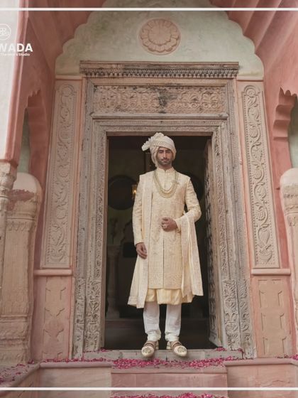 A groom stands at the entrance of a heritage palace, his Shauhar sherwani fitting the scene perfectly. This is the majestic art I strive to create for every modern groom.