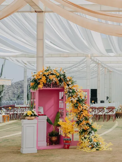 A quirky and fun photo booth for a Haldi ceremony, featuring a pink British-style phone booth overflowing with yellow and orange flowers.