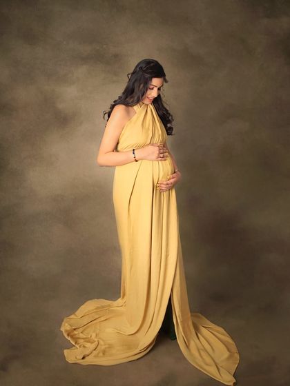 A quiet, intimate moment of connection. This portrait captures a mom-to-be gently cradling her bump, dressed in a beautiful golden halter gown against a soft, textured background.