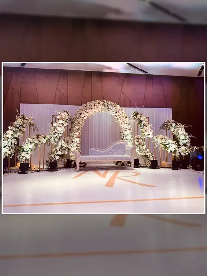 An elegant and sophisticated stage design featuring a stunning arch of white florals. The clean lines, monogrammed floor, and classic white chaise lounge create a timeless and chic backdrop for any celebration.