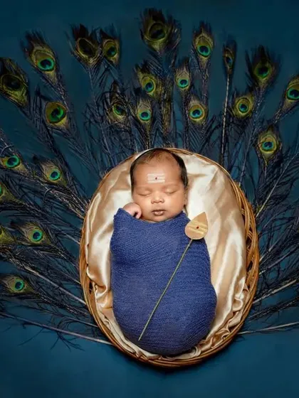 A striking composition with a baby dressed as little Muruga against a deep teal background, framed by a beautiful spread of peacock feathers.