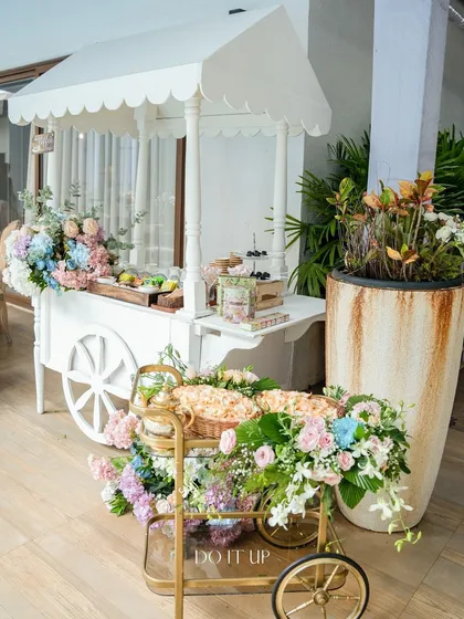 A charming white cart used as a dessert and snack station for a baby shower. It's decorated with pastel floral arrangements, adding a touch of whimsy and elegance to the celebration.