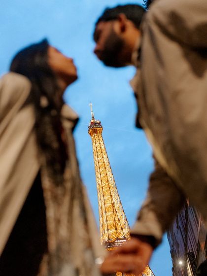 A creative, low-angle shot looking up at the couple with the Eiffel Tower's spire between them. The focus is blurred, creating an artistic and abstract view of a classic Parisian landmark.