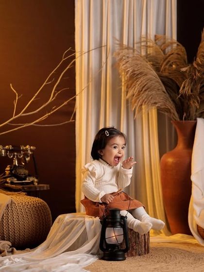 A toddler sits in a rustic, moody setup with dried branches, pampas grass, and vintage props like an old telephone.