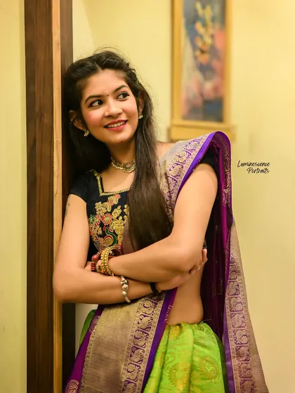 A charming portrait of a woman in a half-saree, her smile radiating warmth as she leans against a wall.