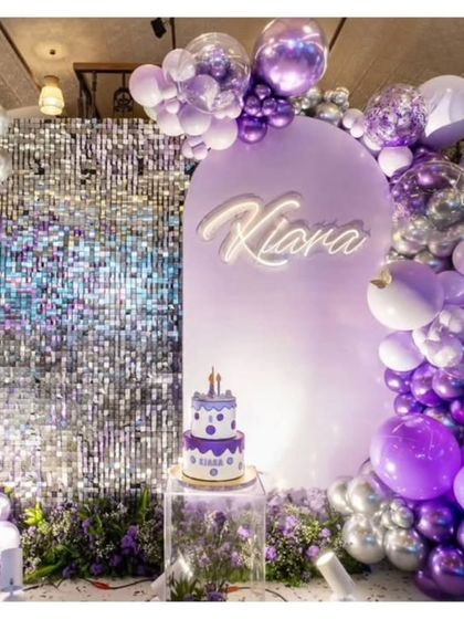 A chic and glamorous birthday setup with a silver sequin shimmer wall and a lavender arched backdrop. The scene is set with a custom neon name sign, a tiered cake on a clear pedestal, and a stunning balloon garland in shades of purple and silver.