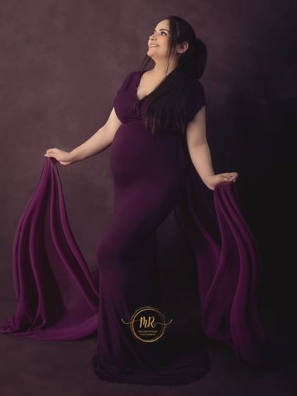 A full-length portrait of an expectant mother in a vibrant purple gown. The flowing fabric trails behind her, creating an elegant and classic maternity look.