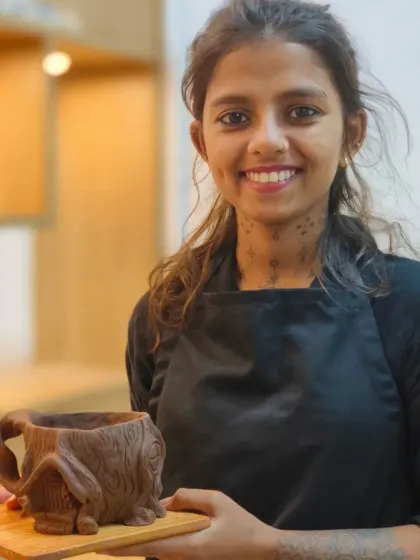 A smiling student holds up her finished tree-bark-textured mug with a house detail. It is a wonderful feeling to complete a piece and see your idea become a tangible object.