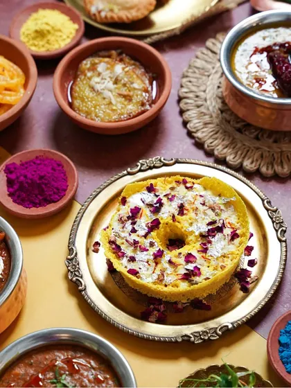 A beautiful Ghevar, a traditional Rajasthani sweet, garnished with rose petals, set on a brass plate for Holi. The surrounding colors and textures make this a rich and festive shot.